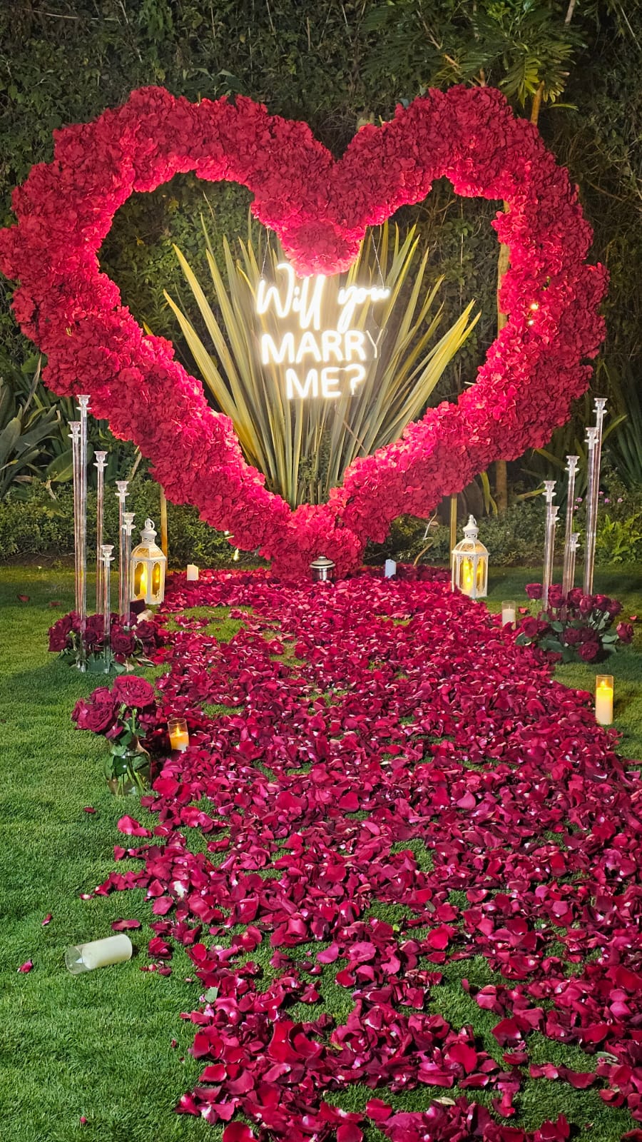 Evening proposal setup with a lit rose heart arch, lanterns, candles and deep red petal aisle