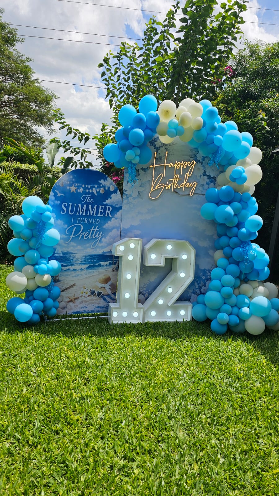 Blue and cream balloon arch with a lit number 12 marquee sign for a garden birthday party