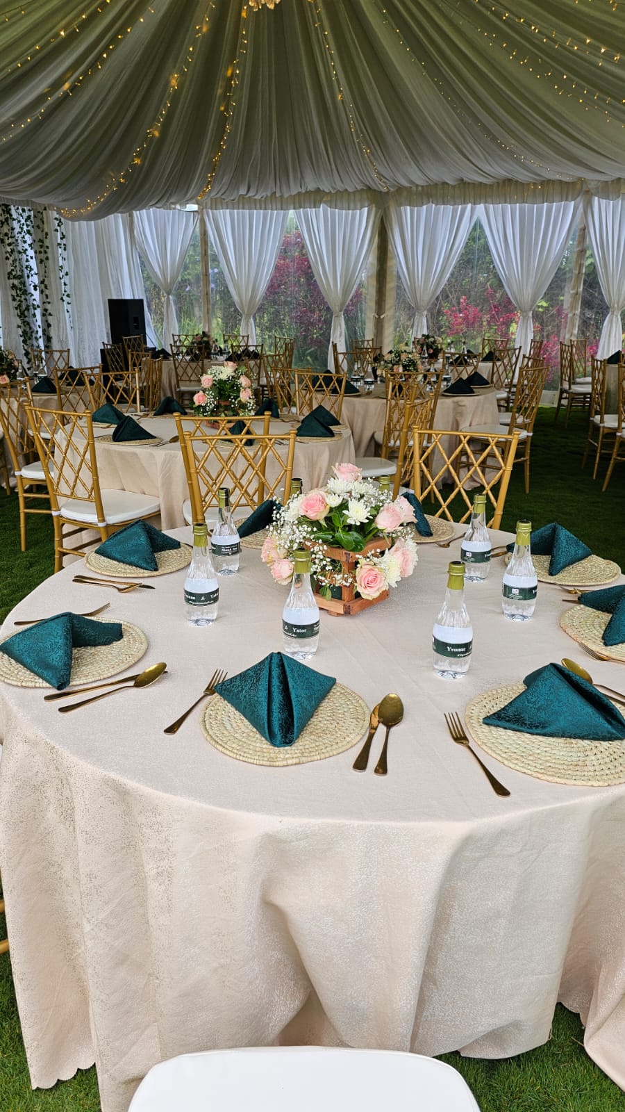 Round reception table with teal napkins, woven placemats and gold cutlery inside a fairy-lit marquee